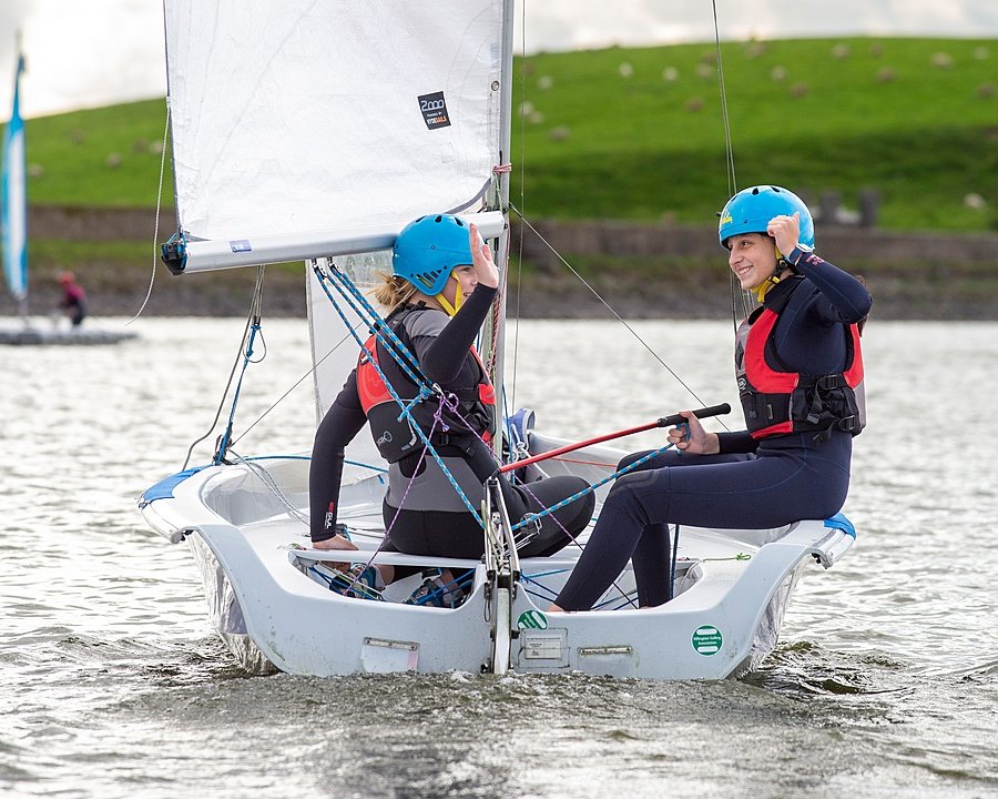 Two pupils sailing a small boat on a lake, wearing helmets and life jackets.