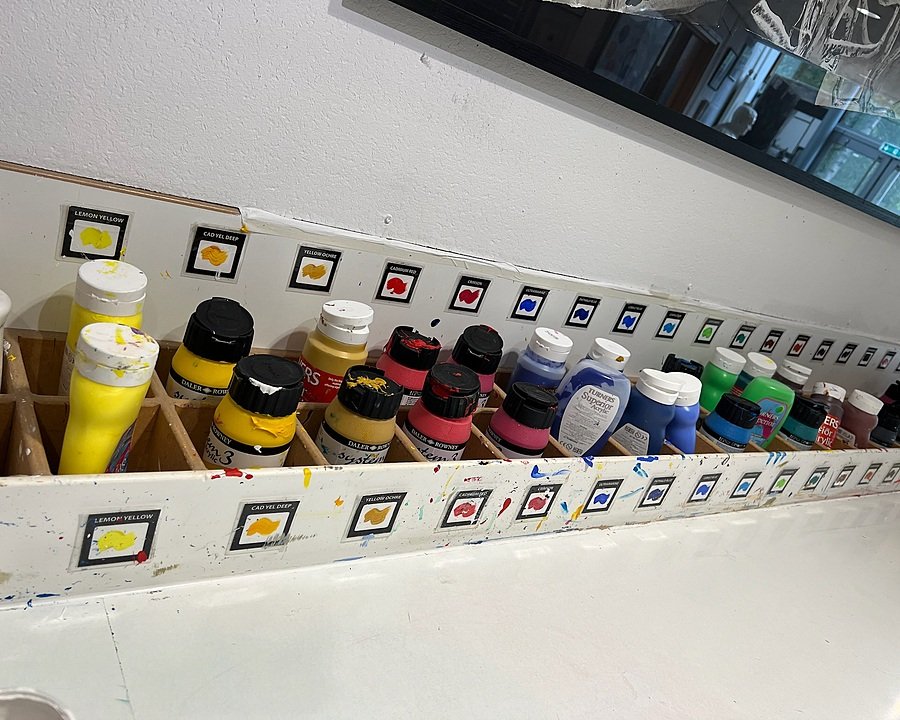 Row of colourful paint bottles neatly organised by shade in an art classroom.