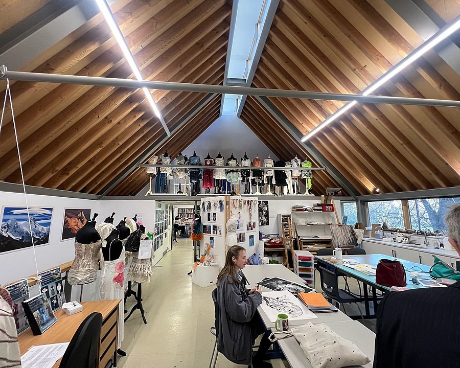Art and design classroom with a high wooden ceiling, mannequins displaying creative garments, and students working at tables surrounded by artwork and fabrics.