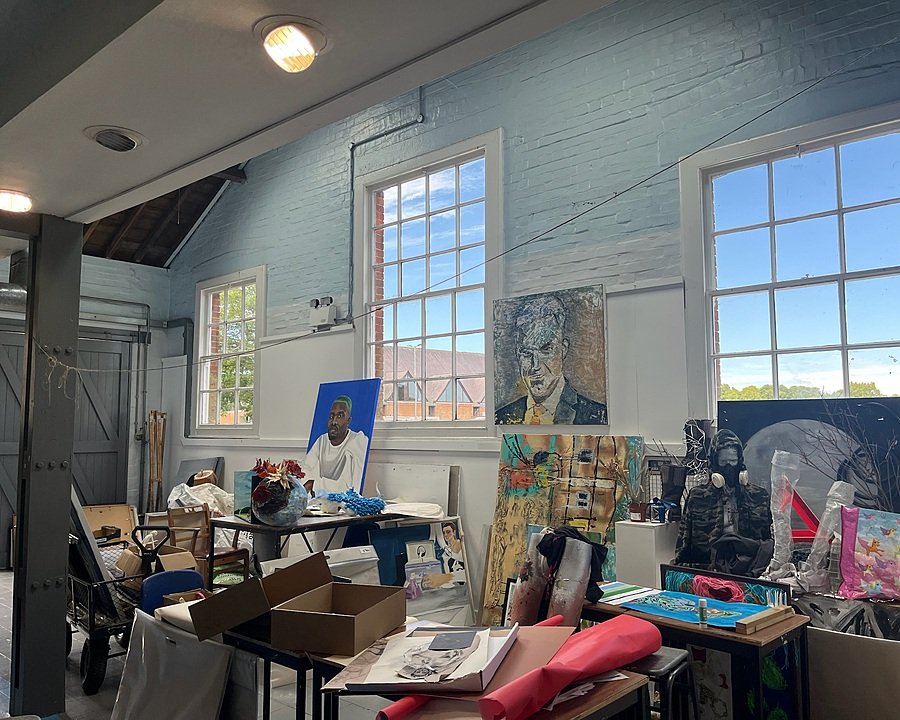 An art studio with large windows and bright natural light, showing various artworks including portraits and abstract paintings displayed around the room.