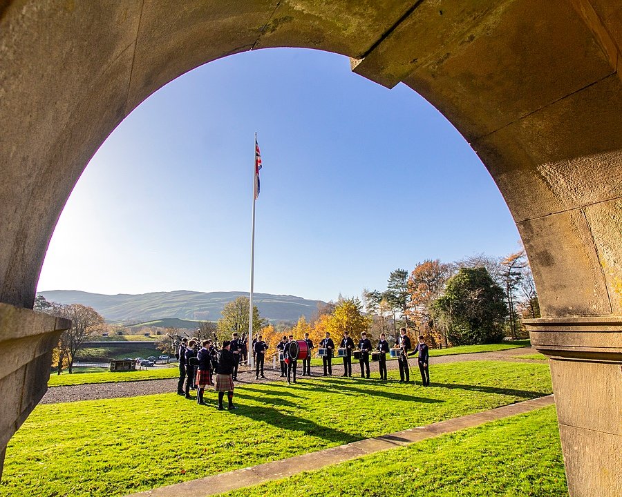 Pupils gathered on a lawn for a flag-raising ceremony under an arch, with hills in the distance.