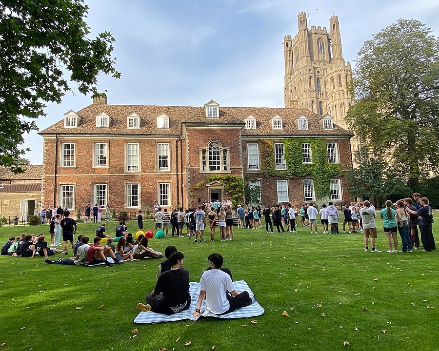 Students gather and relax on a lawn in front of a historic red-brick building covered partly with ivy, with a tall cathedral-like tower in the background.
