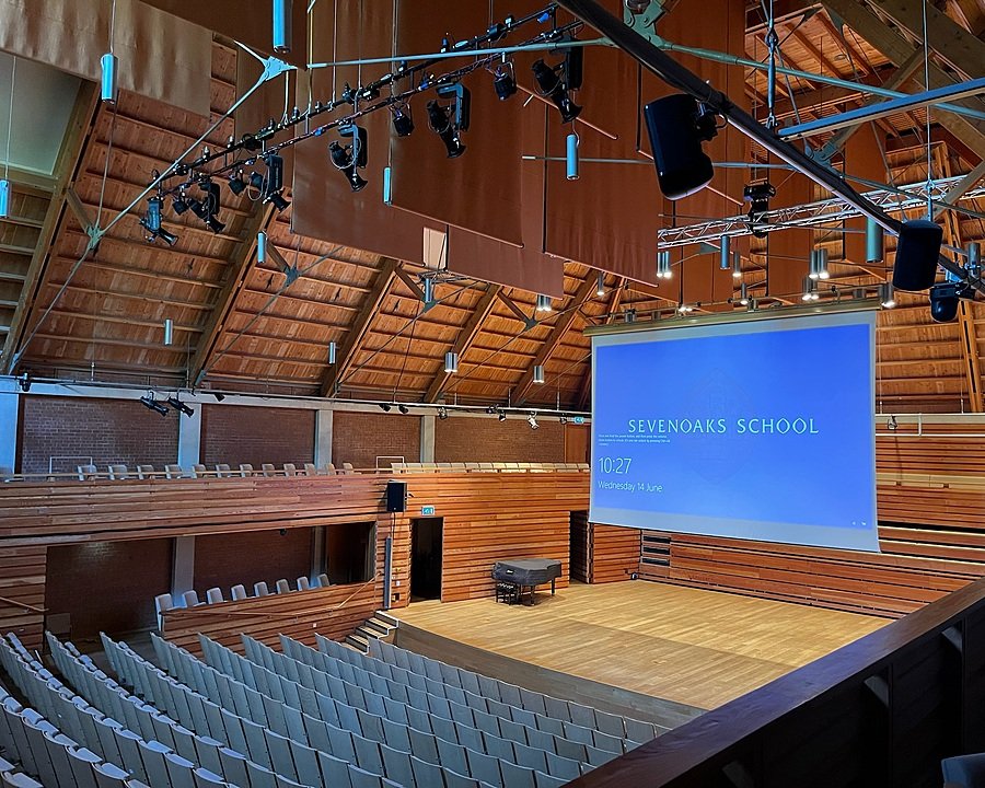 A large wooden auditorium with tiered seating, a stage, and a presentation screen.