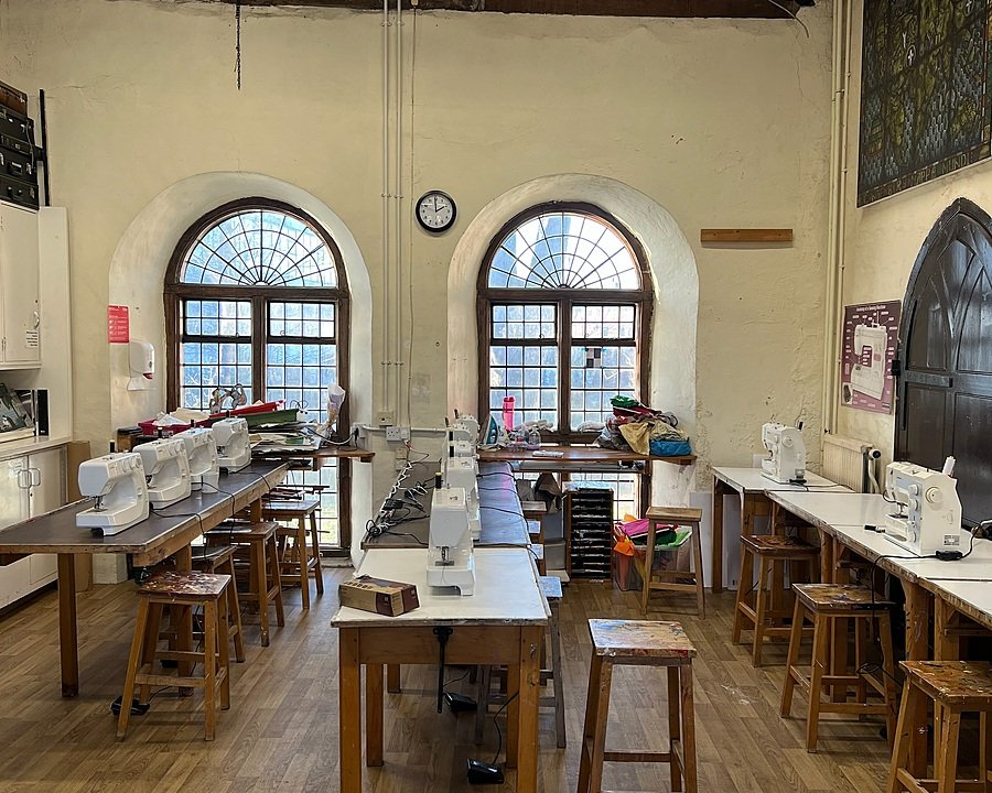 Classroom with sewing machines lined up on wooden tables beneath two arched windows in a historic building.