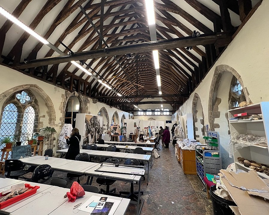 Art studio set in a historic hall with high wooden beams, arched windows, and students working on projects.