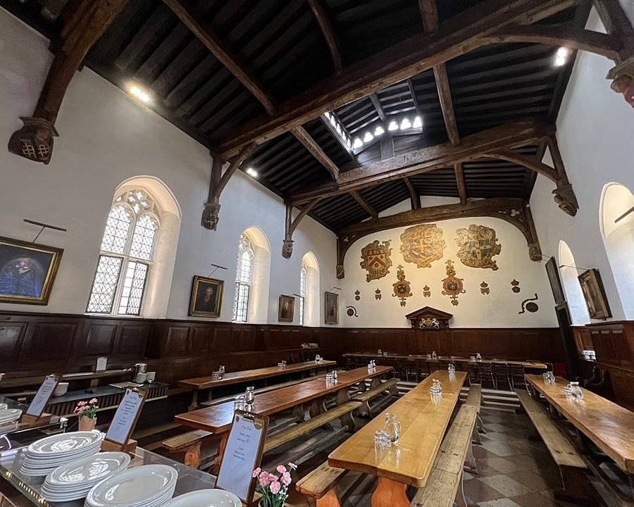 Historic dining hall with wooden beams, long tables, and coats of arms painted on the far wall.