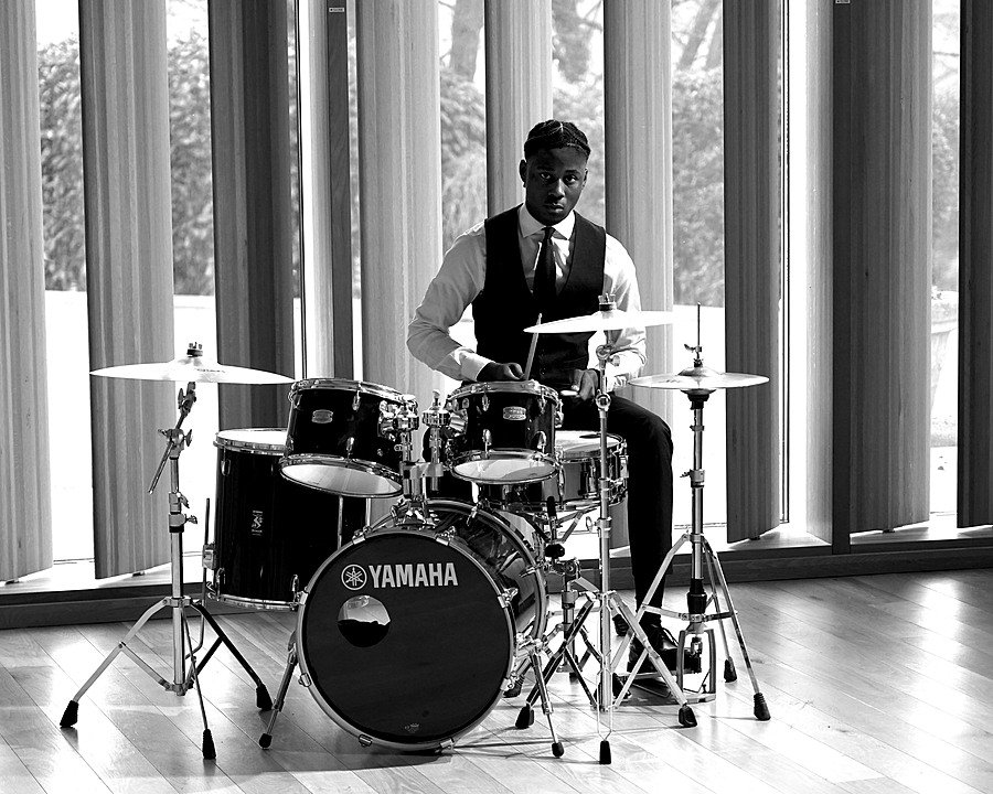 A boy in waistcoat and shirt playing the drums with a modern window front in the background