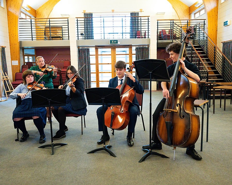 Students play string instruments, including violins, cello, and double bass, during a music rehearsal in a bright hall.