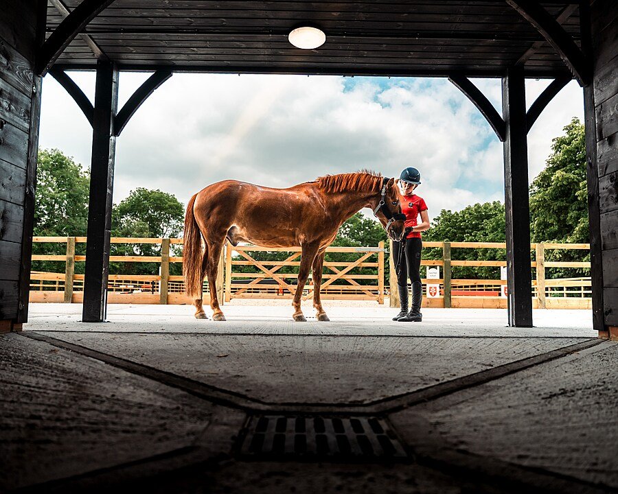 Student leading a horse in a covered stable yard with riding arena behind.