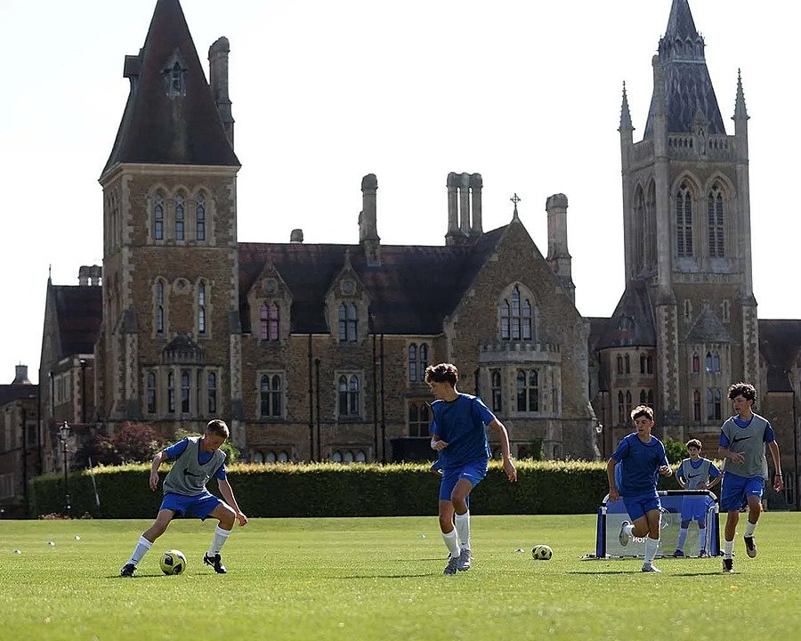 Pupils practising football on a green field with an impressive historic school building in the background.