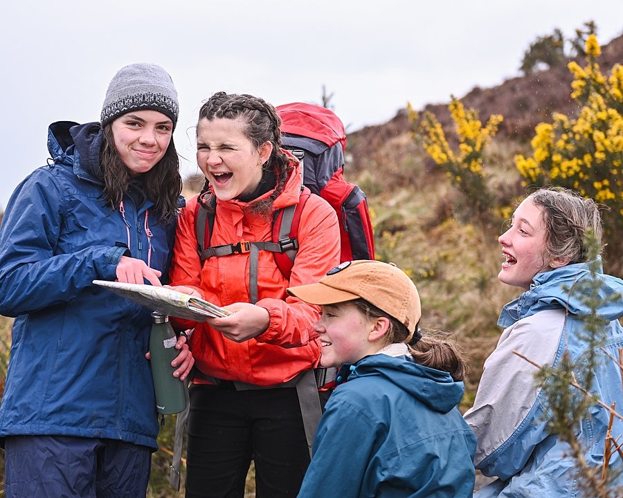 Four girls dressed in waterproof outdoor clothing laugh together while looking at a map during a hike through a hilly, rural area with yellow flowering bushes.