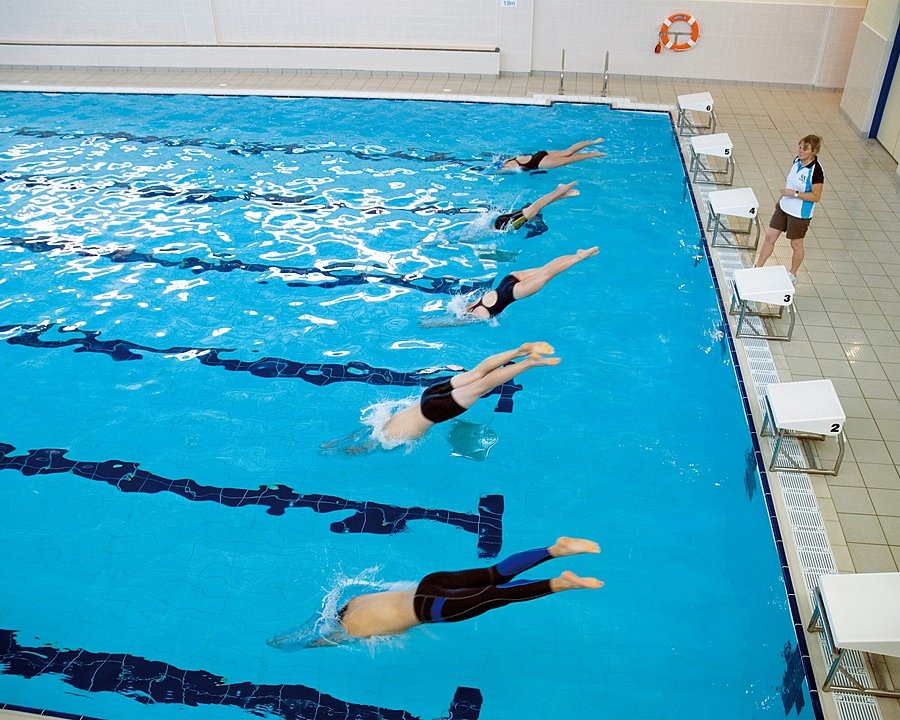 Swimmers diving into a clear blue indoor pool at the start of a race, while a coach watches from the side.