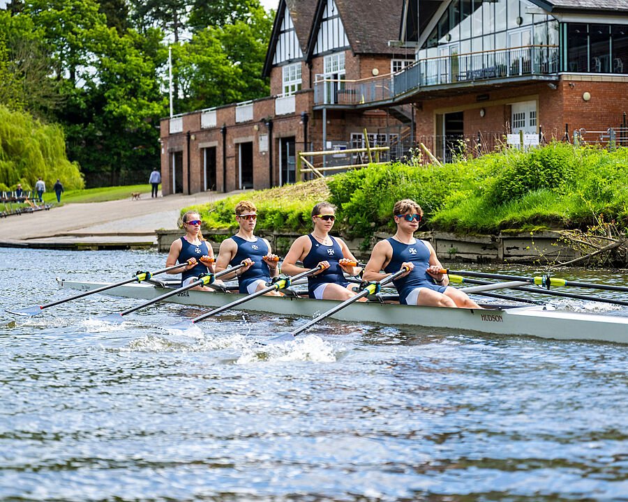 A rowing team gliding across the water near a boathouse on a sunny day.