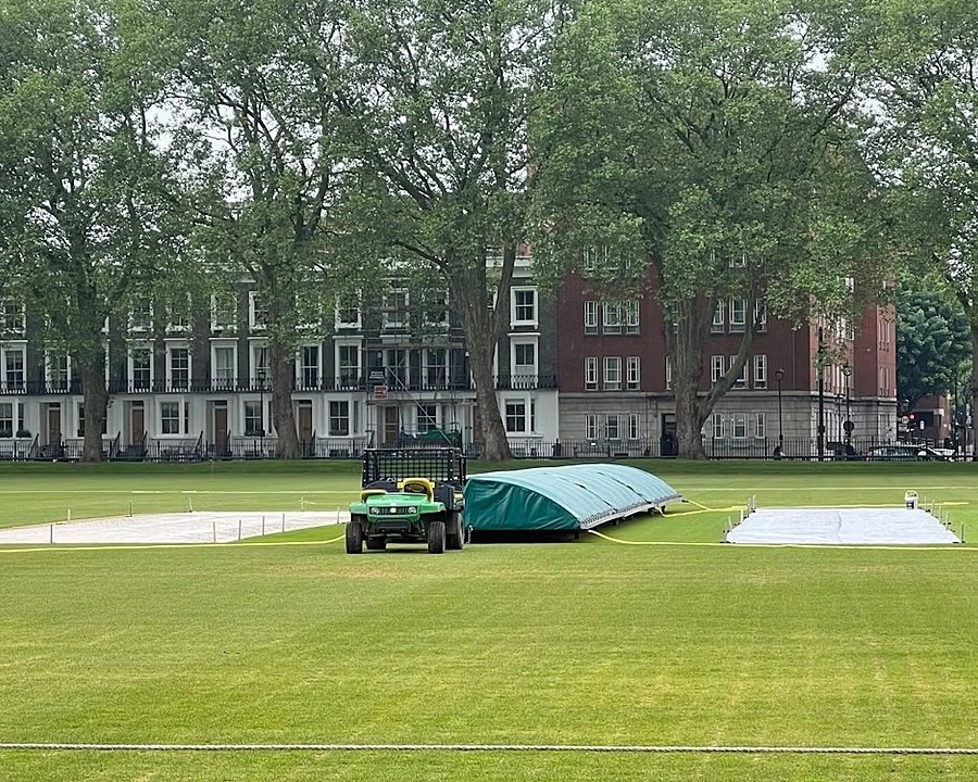 Cricket ground with covers on the pitch, surrounded by tall trees and Georgian-style buildings.