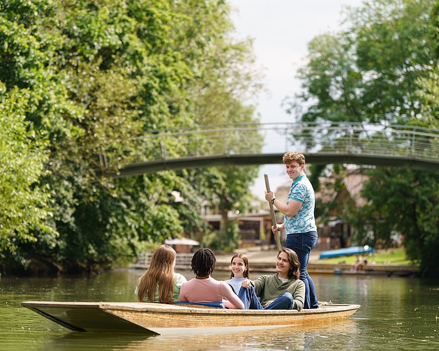 Students punting on a calm river surrounded by lush green trees, with a curved pedestrian bridge in the background.