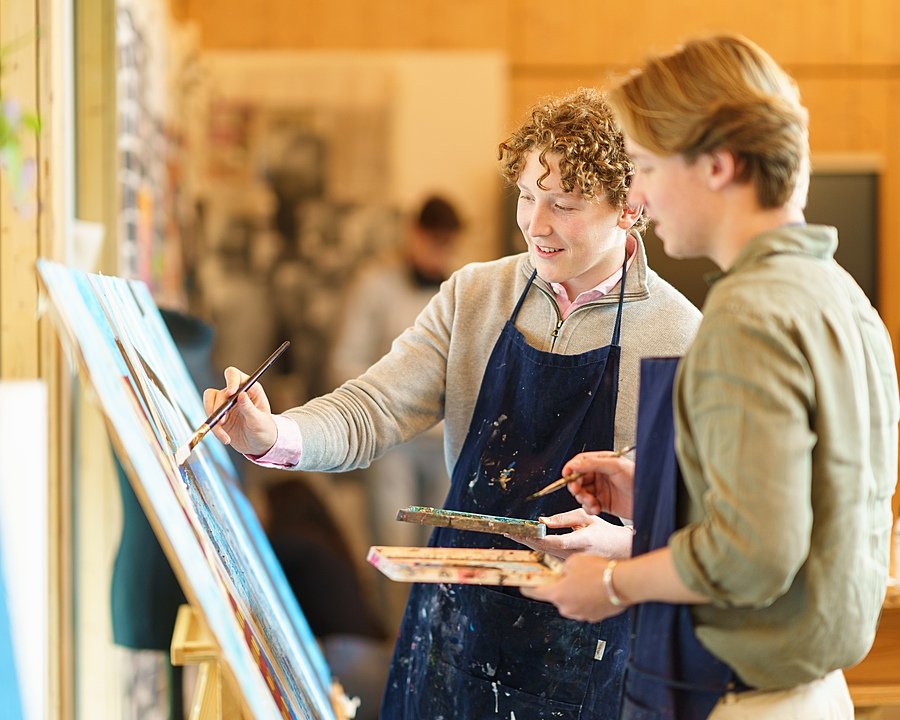 two students in aprons painting with brushes on an easel