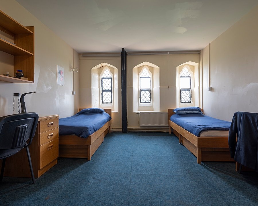 Simple boarding room with two single beds, blue bedding, wooden desks, and arched windows letting in natural light.