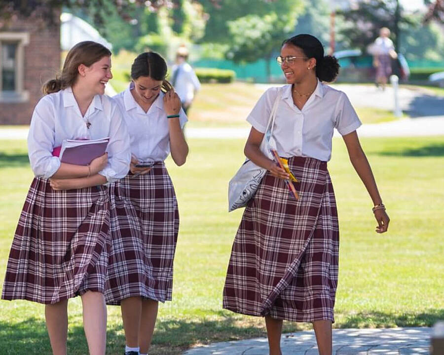 Three girls in school uniforms with white shirts and plaid skirts walking and laughing together on a sunny day.