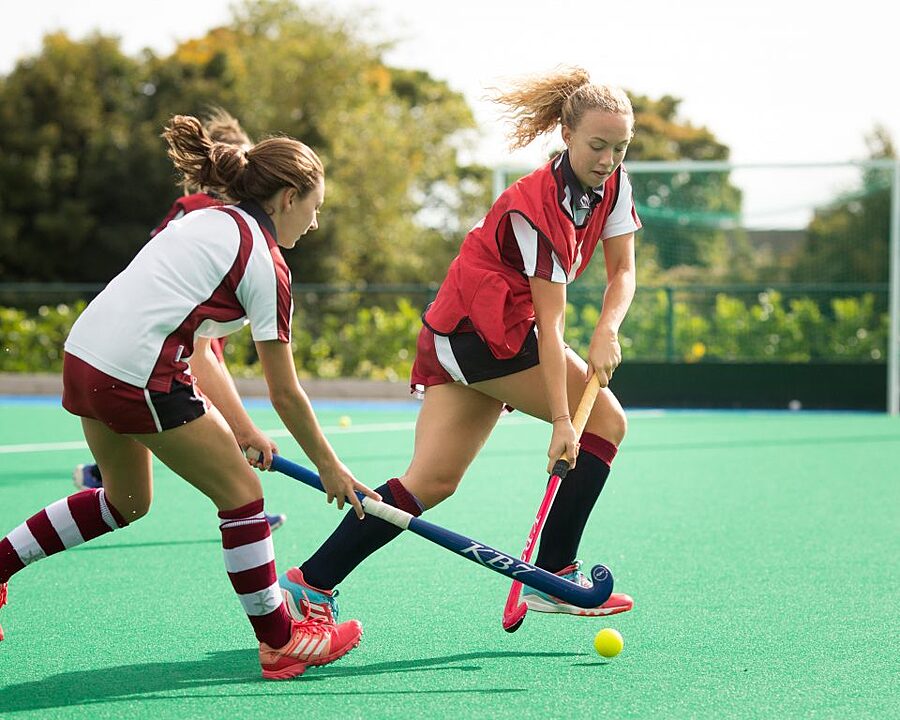 Two girls in hockey gear playing on an artificial turf field; one is dribbling the ball while the other tries to defend.