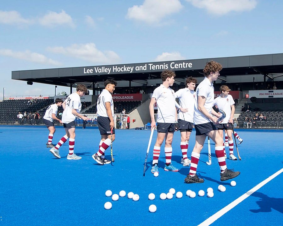 Group of boys in white and blue hockey uniforms standing on a blue hockey field with white balls, at the Lee Valley Hockey and Tennis Centre.