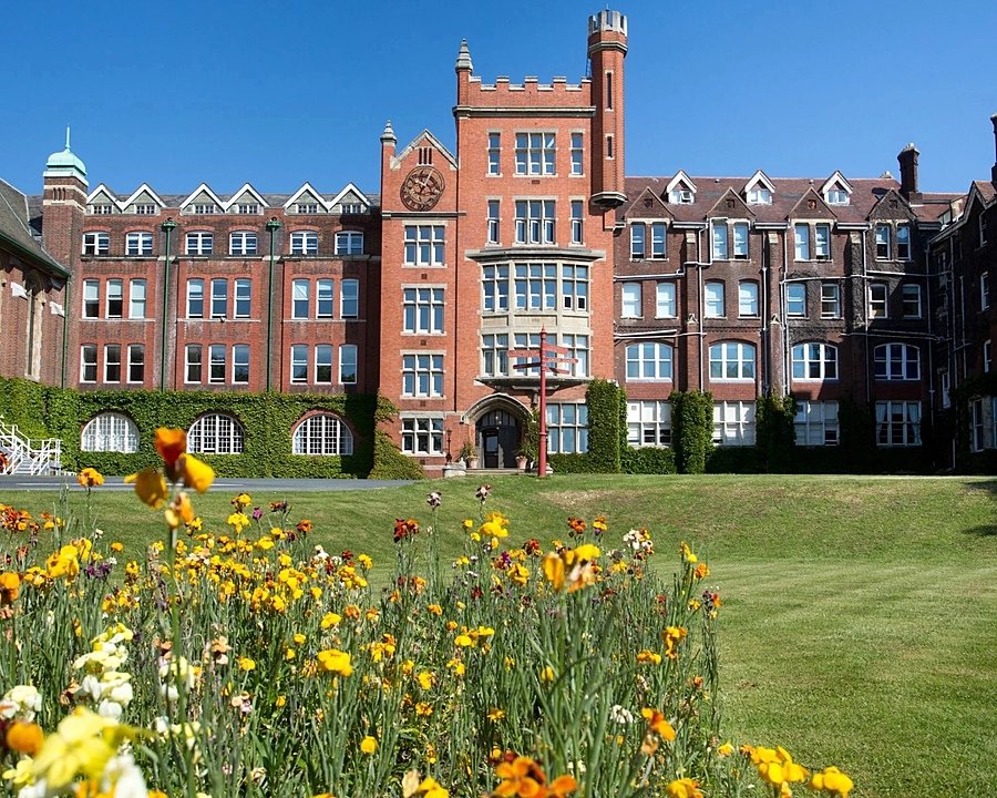 Large historic red-brick school building with a clock tower, seen from a lawn with colorful flowers in the foreground under a clear blue sky.