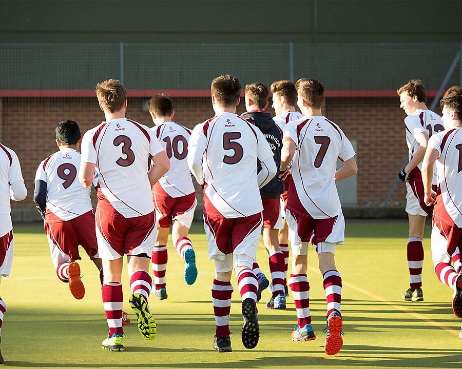 Group of boys in white and maroon sports uniforms running together on a hockey field during training.