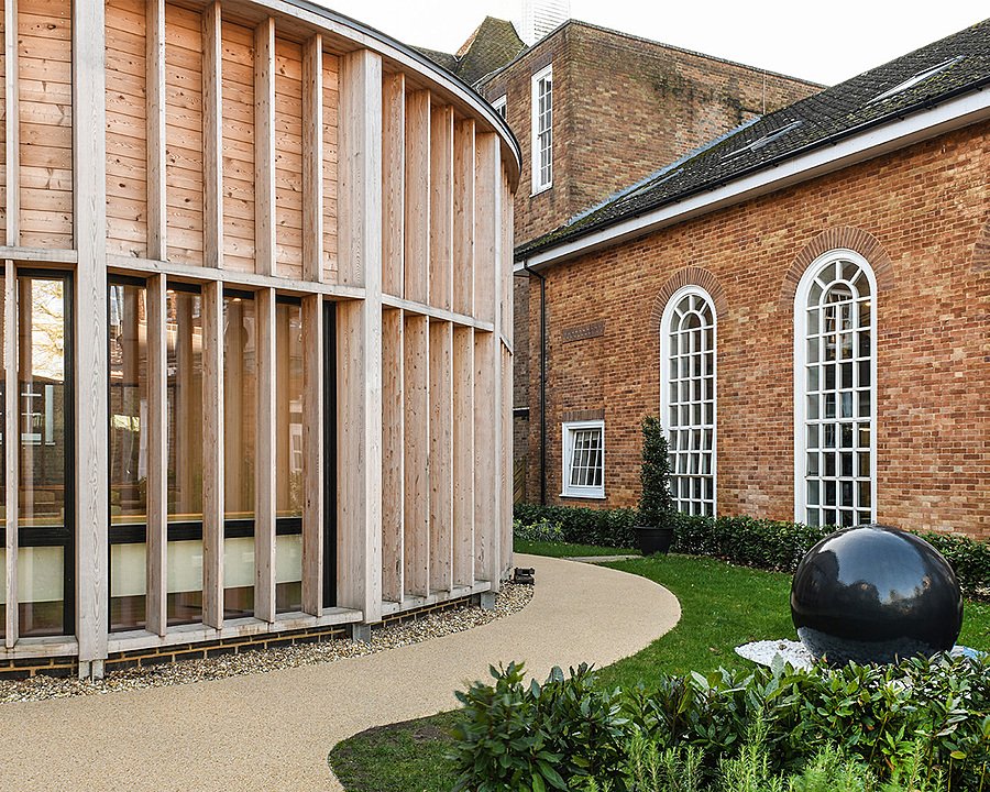 A modern, round, wood-panelled building with large windows next to a red brick building