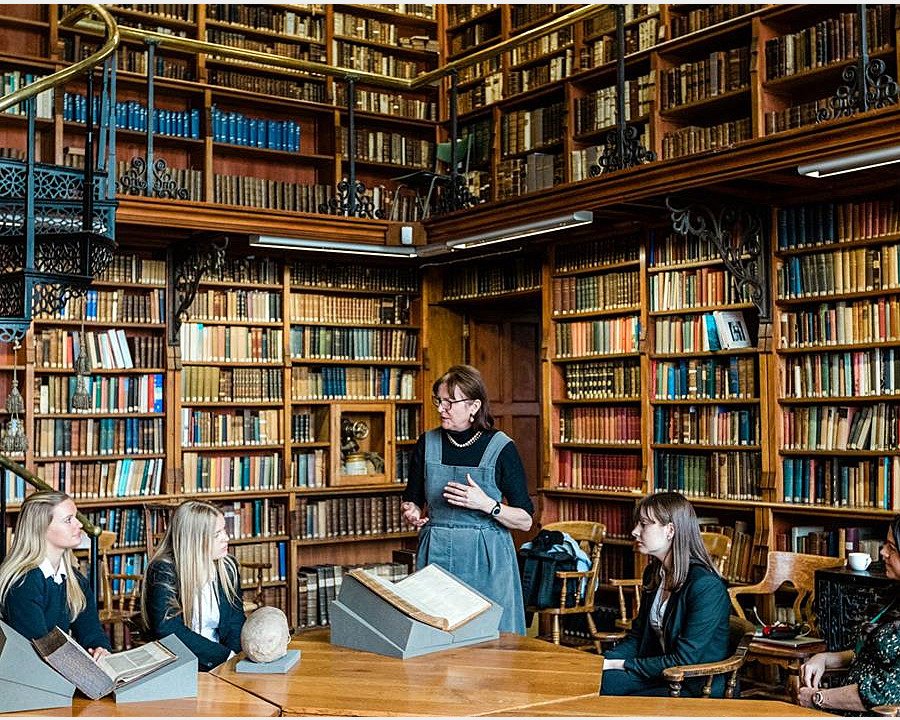 a teacher with her students in an old library with books up to the ceiling and iron spiral stairs leading up