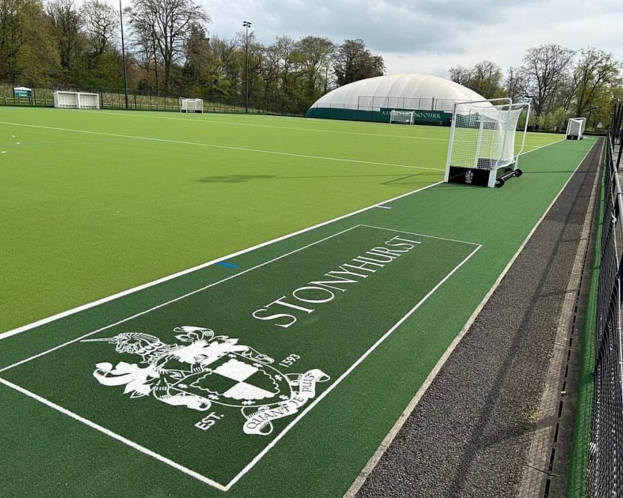 A green artificial hockey field with white goals and “Stonyhurst” written beside the crest near the sideline.