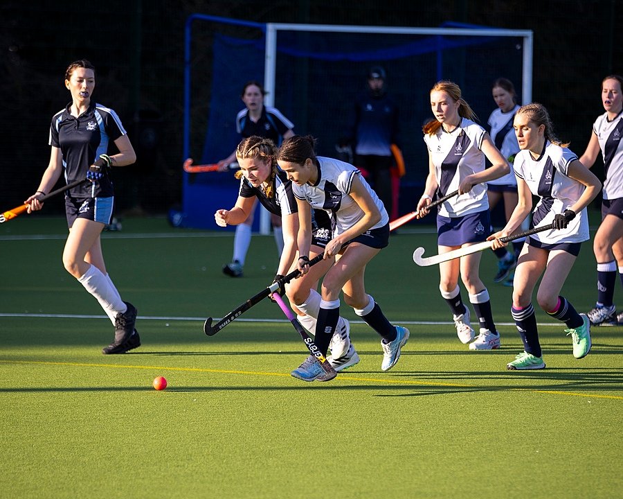 Girls playing field hockey on an outdoor pitch in sports uniforms during a match.