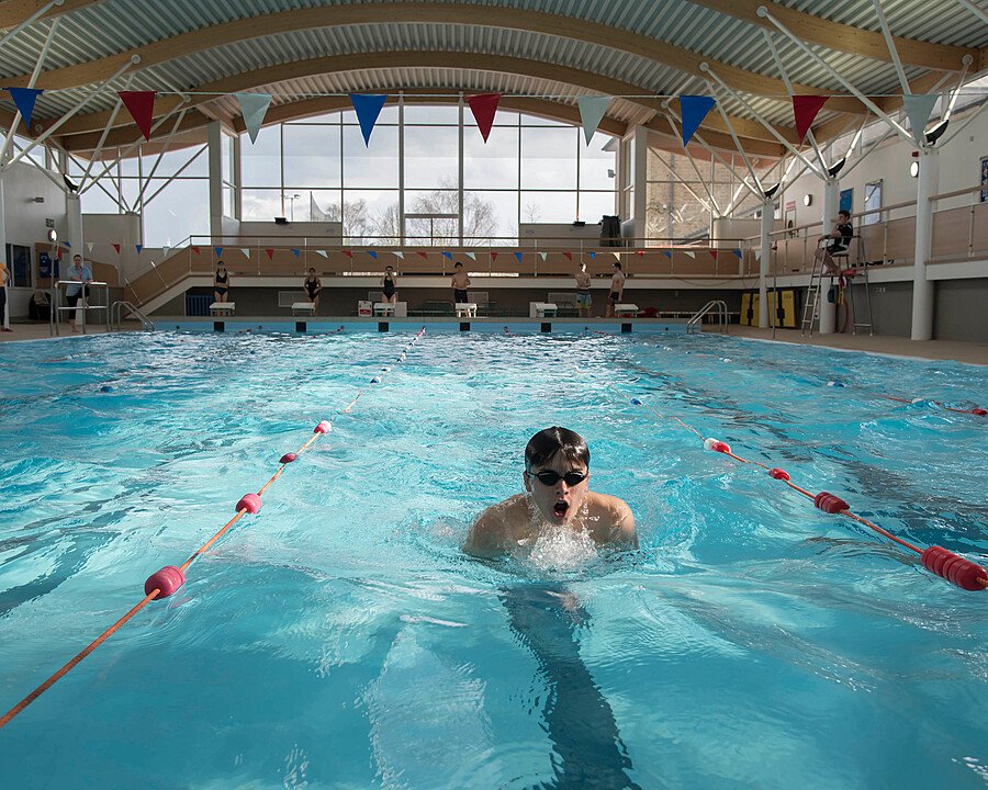 Swimmer training in an indoor swimming pool with lifeguards and large windows in the background.