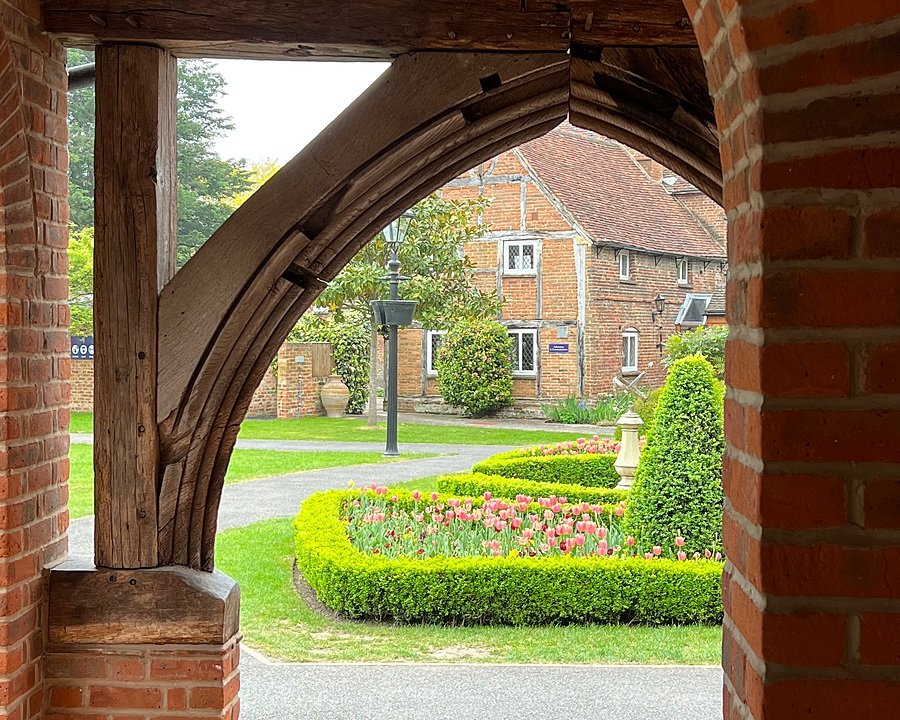 View through a brick and timber archway onto a courtyard with neatly trimmed hedges, tulip flower beds, and an old brick building in the background