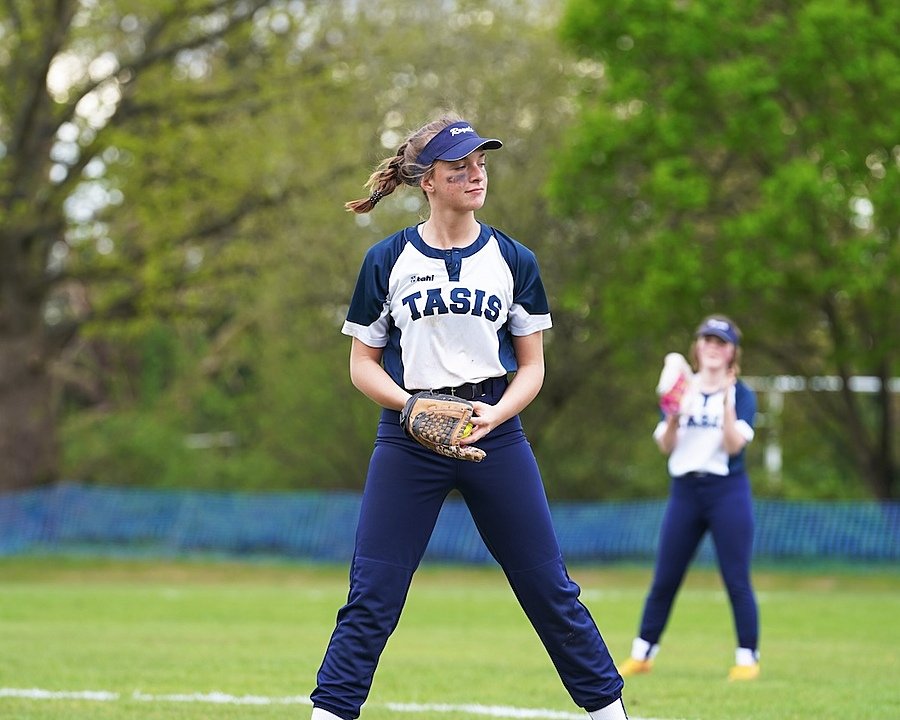 Girl in a TASIS sports uniform playing softball on a grassy field, with teammates and a coach in the background