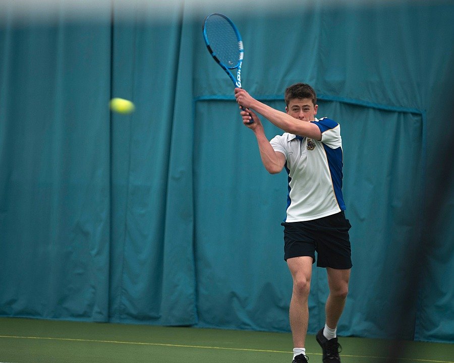 Student playing indoor tennis, preparing to hit a forehand shot against a blue curtain backdrop.
