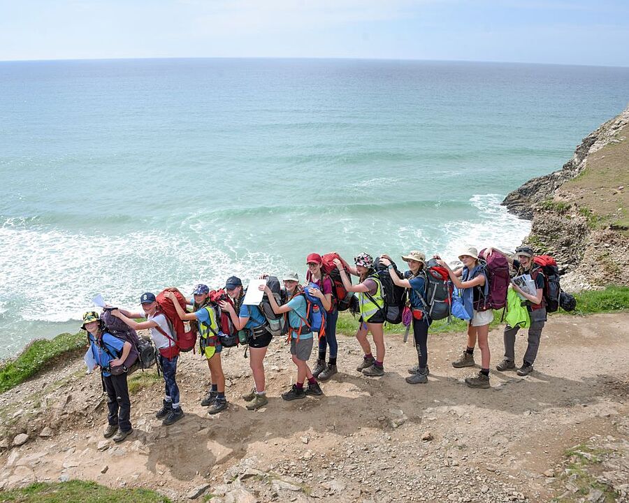 Group of students with large backpacks standing on a cliffside path overlooking the ocean.