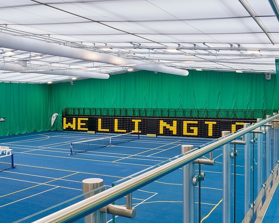 Large indoor tennis facility with blue courts, green curtains, and bright overhead lighting.