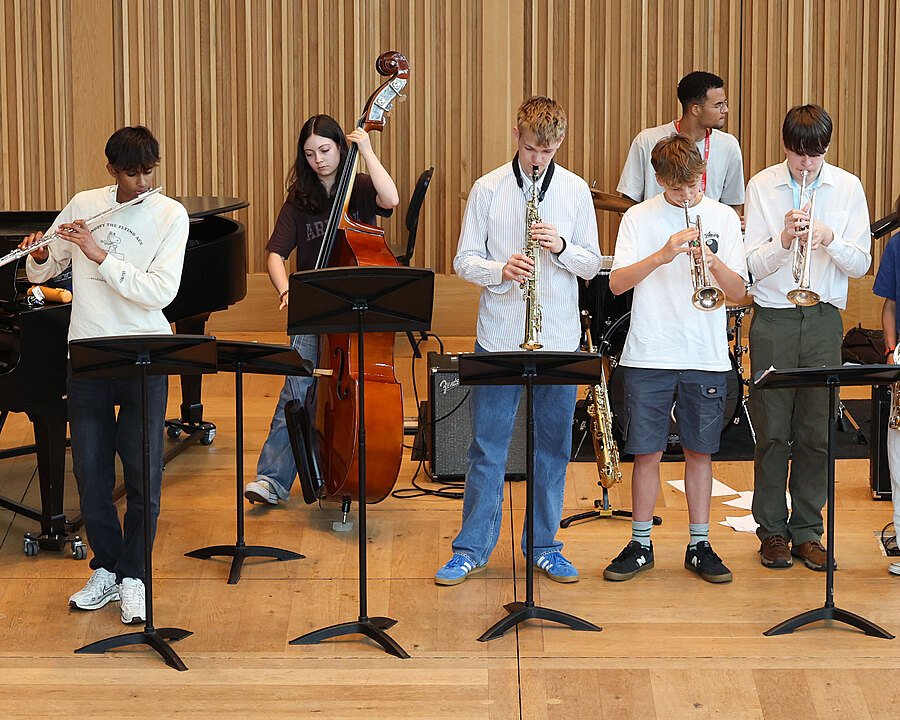 A group of students playing instruments including trumpet, saxophone, and piano during a music rehearsal on a wooden stage.