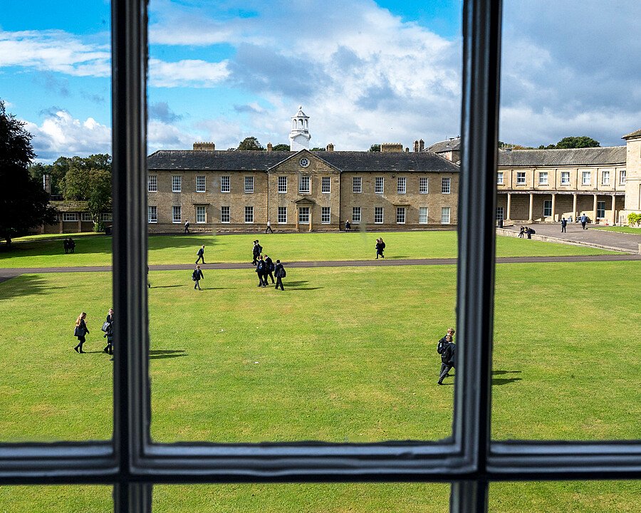 A view from a window overlooking a school courtyard where students walk across a large grassy area surrounded by stone buildings.