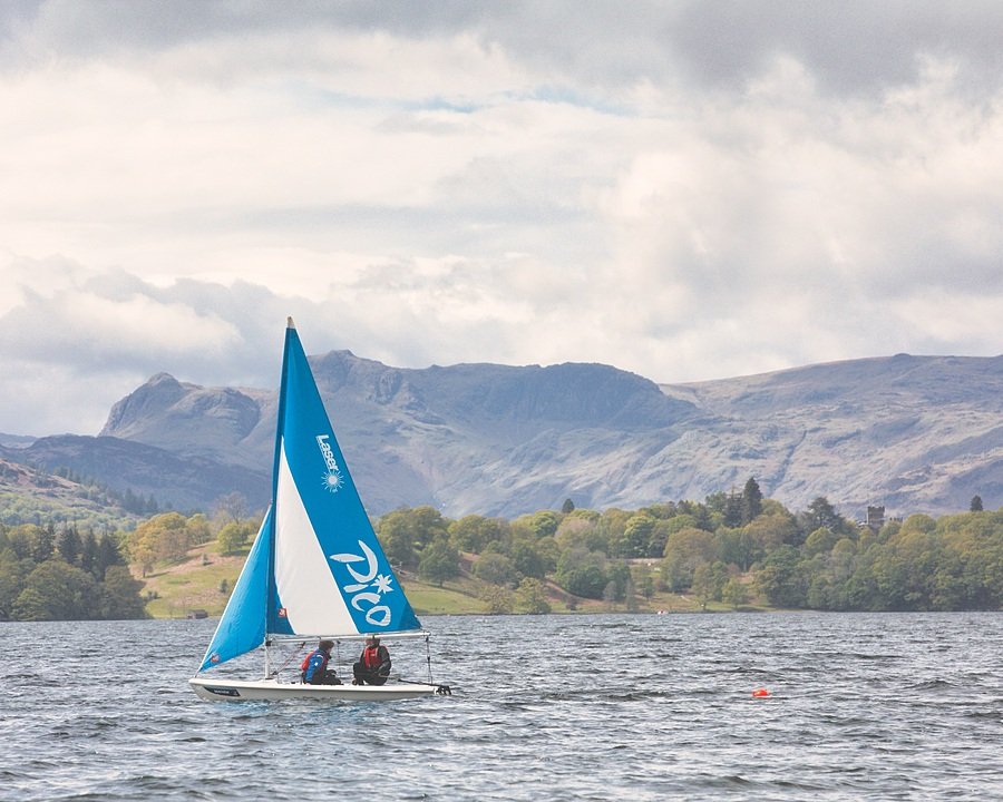 Two students sailing a small dinghy on Lake Windermere with the Lake District mountains in the background.