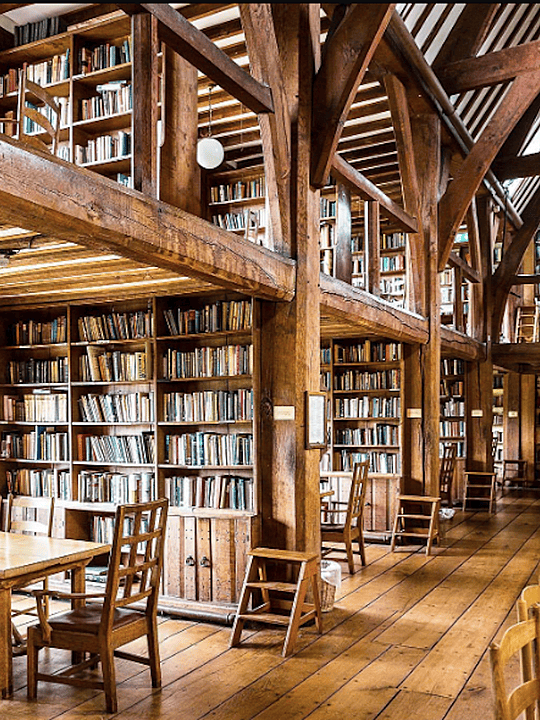 An interior view of a large, historic library with wooden beams, shelves full of books, and wooden tables and chairs for studying.