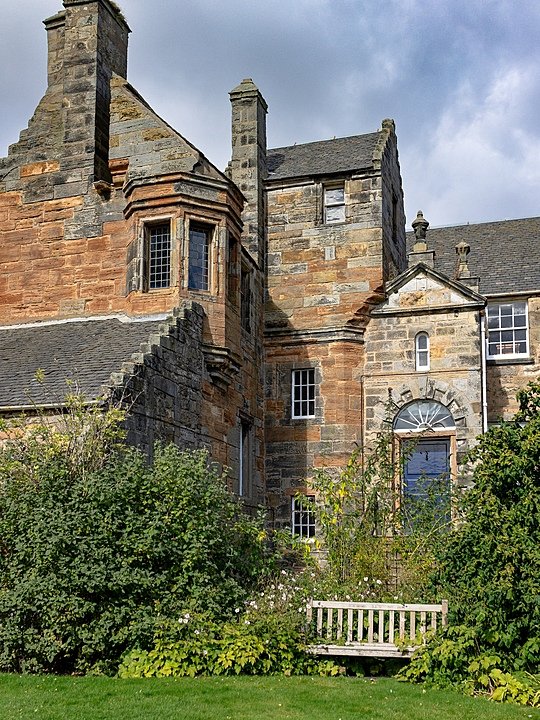 Stone building with historic architecture, featuring tall chimneys and a bay window surrounded by greenery.