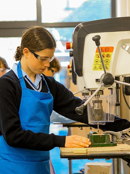 Student wearing safety glasses and a blue apron operating a drill press in a workshop.