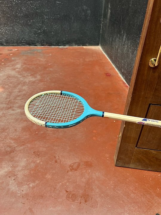 Close-up of a wooden racquet held over a red court floor, likely used for an indoor racket sport.