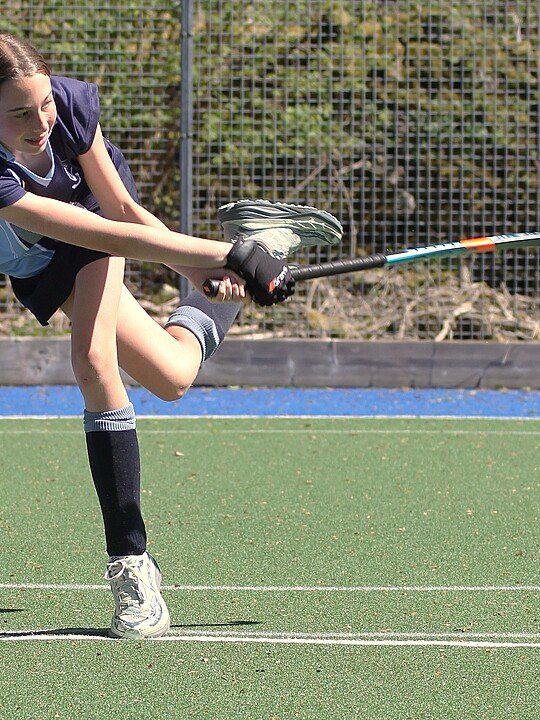 Student striking a hockey ball during training on an artificial pitch.