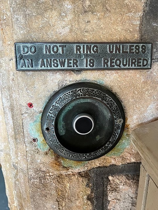 A round metal doorbell with decorative engravings mounted on stone, with a sign reading “DO NOT RING UNLESS AN ANSWER IS REQUIRED.”