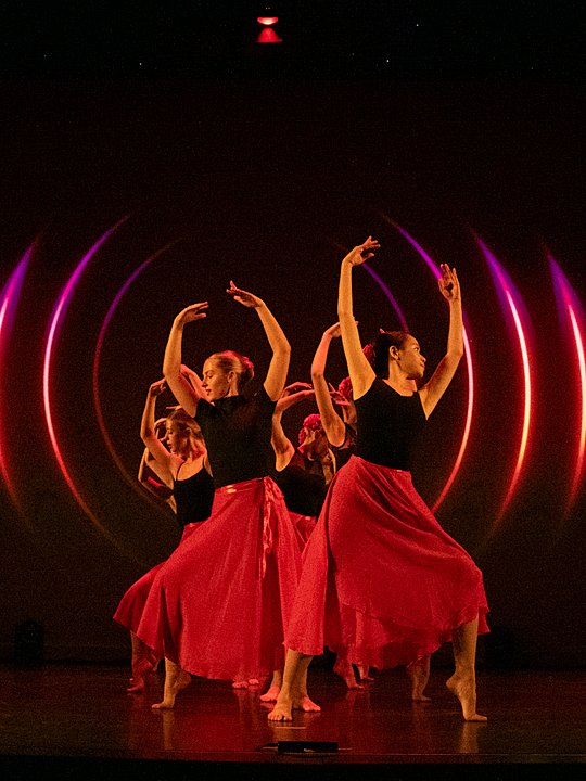 Students performing a choreographed dance on stage with red lighting and flowing costumes.