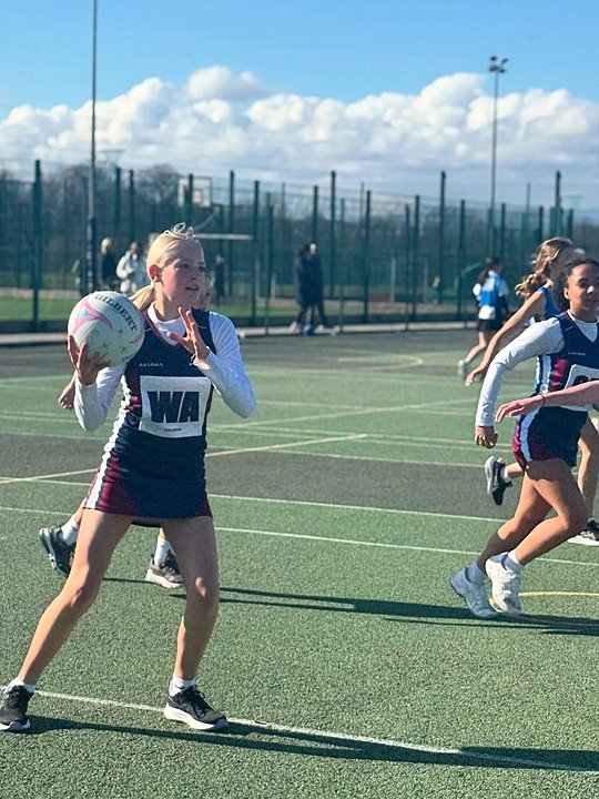 A girl is playing netball on an outdoor court while other players run in the background. She is wearing a school sports uniform with the letters “WA” on her bib.