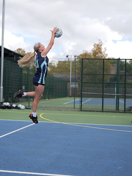 Student in sports uniform jumping to catch a netball on an outdoor court, with tennis courts and trees visible in the background.