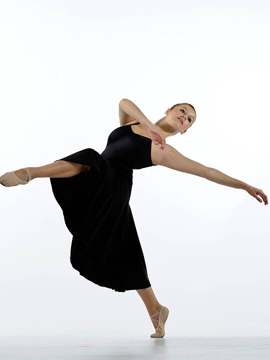 A ballet dancer in a black dress performs a graceful pose on one foot against a white background.