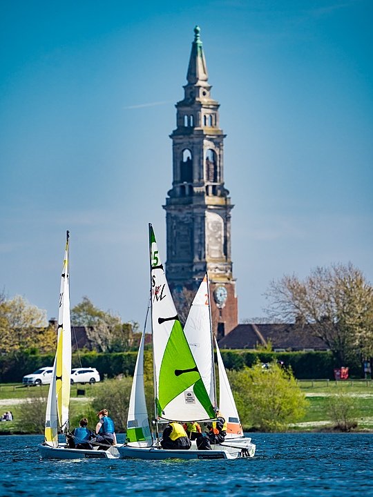 Pupils sailing small boats on a calm lake with the school’s tall clock tower rising in the background.